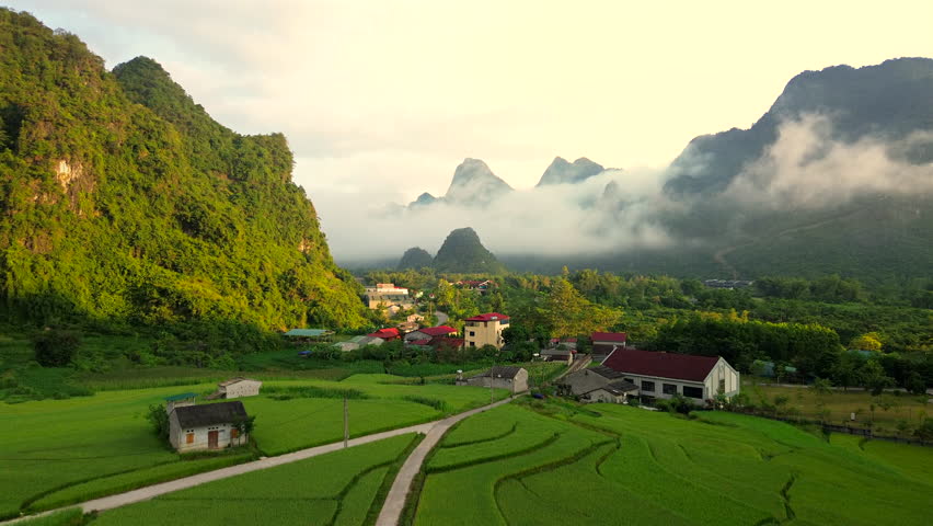 Stunning drone shot of a rural Vietnamese village nestled between limestone karst mountains and green rice fields under a misty morning sky.