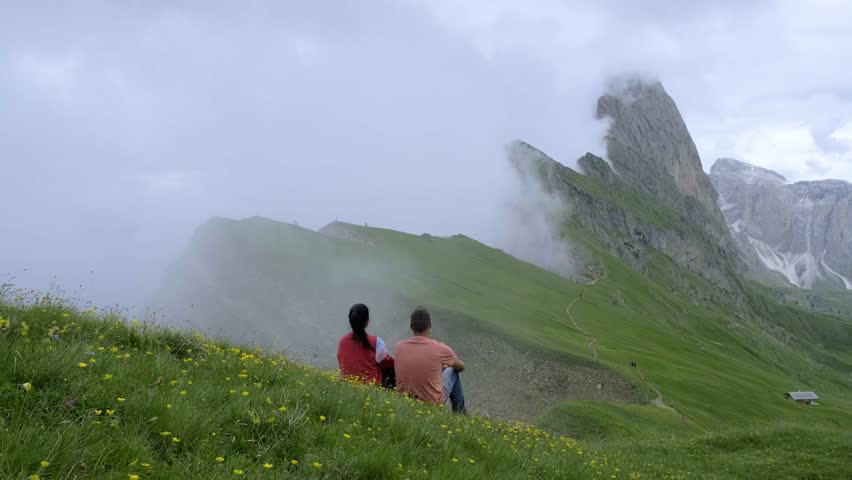 Couple enjoys a serene moment together in the breathtaking Seceda Dolomites, surrounded by lush greenery and majestic peaks under a cloudy sky, evoking a sense of peace and connection to nature.