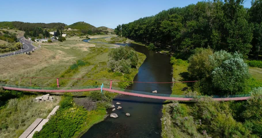 Drone aerial landscape of a winding river with a small footbridge, green riverbanks and dense trees, with hills and open countryside in the background under a clear blue sky.