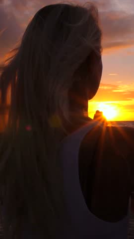 Blonde woman silhouette on a windswept beach at golden hour, long hair trailing as she watches the orange sea and sky, a calm, contemplative sunset moment