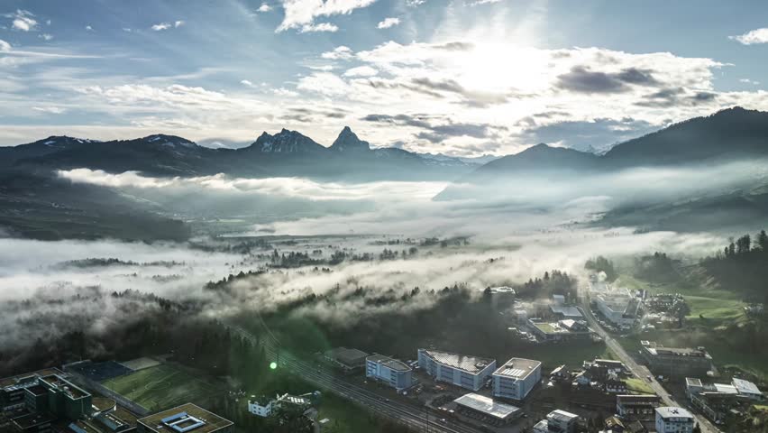 Aerial view looking down at a town with industrial and commercial buildings situated in a valley, with a dense layer of low fog weaving through the landscape and mountains in the distance.