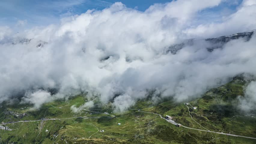 Dramatic aerial view of a serpentine mountain pass road partially obscured by rapidly moving heavy fog and clouds in the Swiss Alps. A moody and atmospheric travel scene.