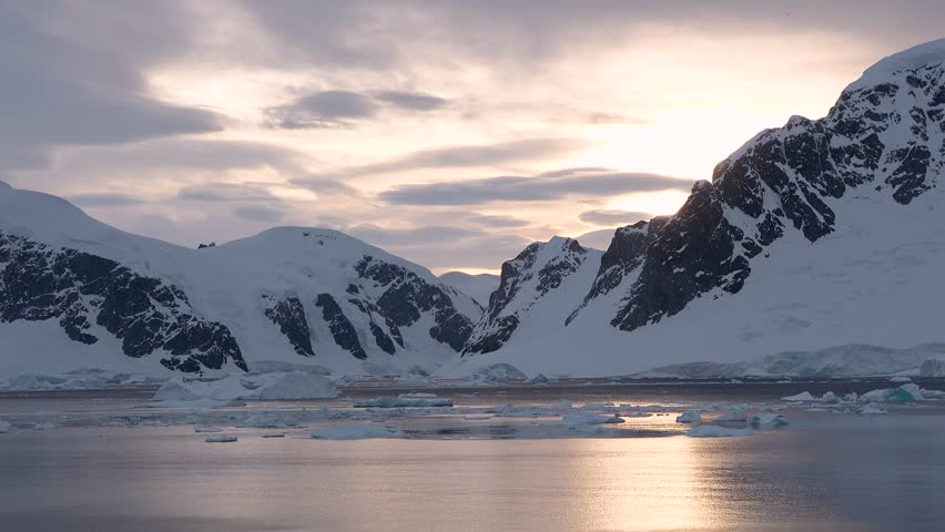 Icebergs drift in icy water as tall mountains stand in the background. The sky is cloudy, and the landscape shows the beauty of Antarctica on a cold day.