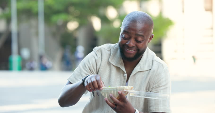 African American man in short-sleeve shirt opening clamshell container, lifting sandwich to eat. Plaza, urban, outdoor, candid, lunch, greens, watch