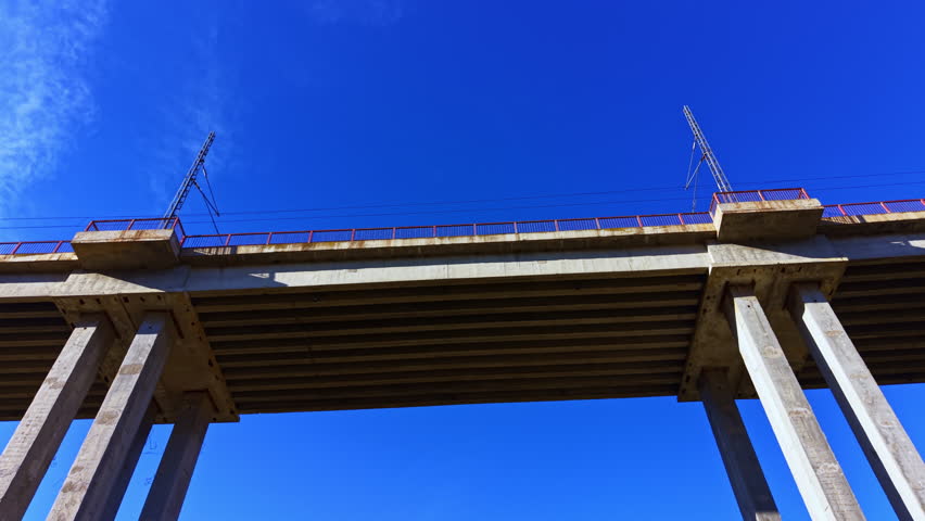 Concrete bridge seen from below with support columns and metal tracks. Bright blue sky provides a backdrop. Scene shows engineering and construction work in an urban setting.