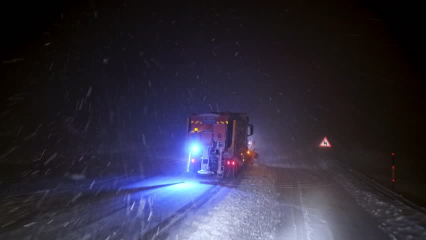 Interior driver view following a snowplow truck clearing a snow-covered road at night. Flashing warning lights, blowing snow and low visibility capture dangerous winter driving conditions.