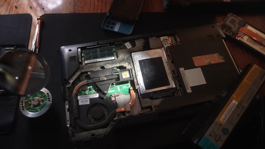 A close up view of a technician wearing gloves working on the internal components of a disassembled laptop, showing components under a magnifying glass. Slow motion.