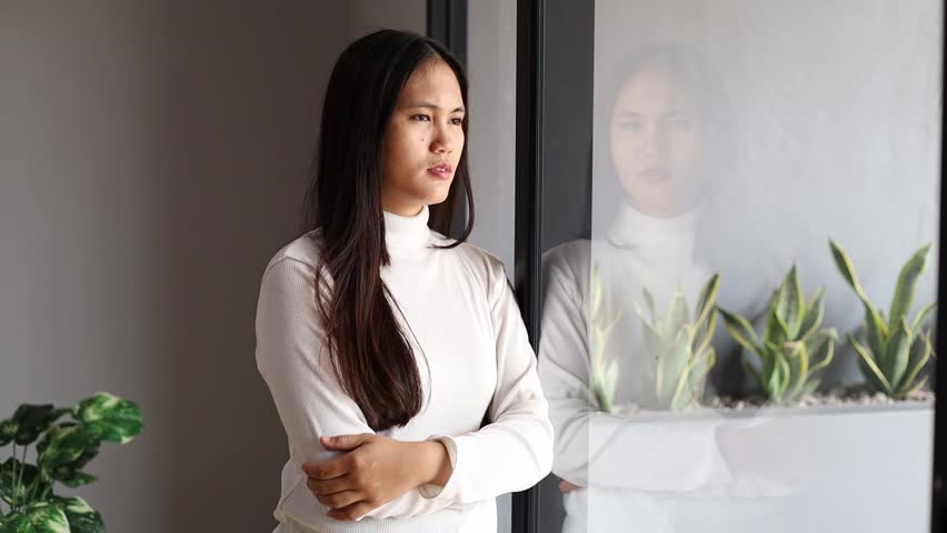 Asian woman with crossed arms pensively through window at home