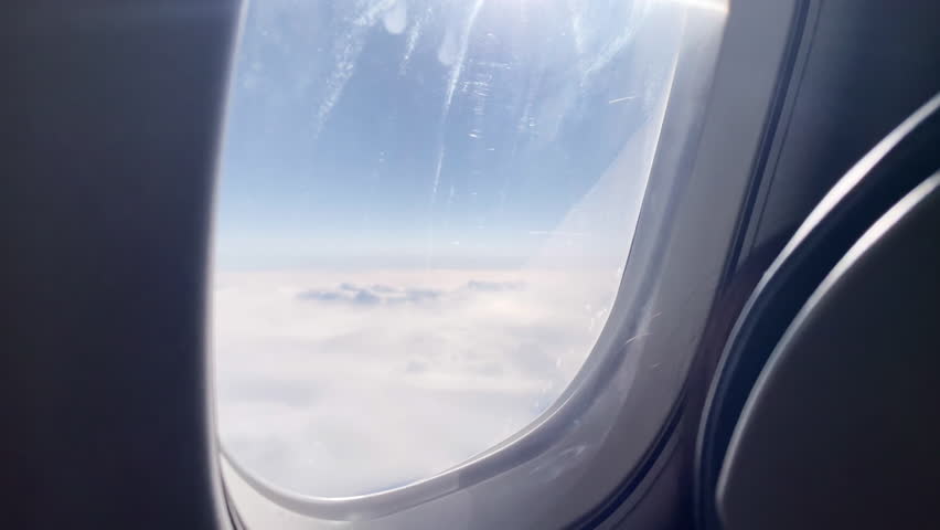 Airplane Window View Over Clouds, Travel Flight Window Seat Perspective. A peaceful airplane window view showing soft clouds and blue sky from a flight window seat, evoking travel, wanderlust, calm and aerial perspective during a daytime journey above the clouds.