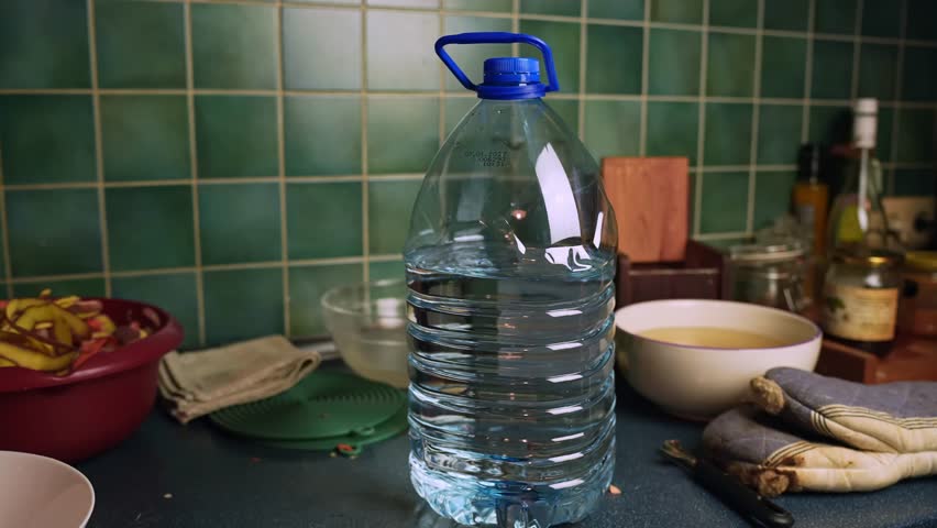 Large plastic water jug with handle standing on a kitchen counter against green tiled backsplash, with bowls and cloths around. Home cooking, hydration, and household routine scene.