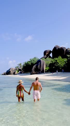 A couple enjoys a romantic walk hand in hand along the crystal-clear waters of La Digue, Seychelles. The beautiful granite rocks and lush greenery create a perfect tropical paradise.