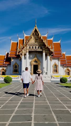 A couple walks hand in hand to the stunning Wat Benchamabophit, the Marble Temple, embraced by vibrant architecture and a clear blue sky, embodying travel romance. Bangkok Thailand.