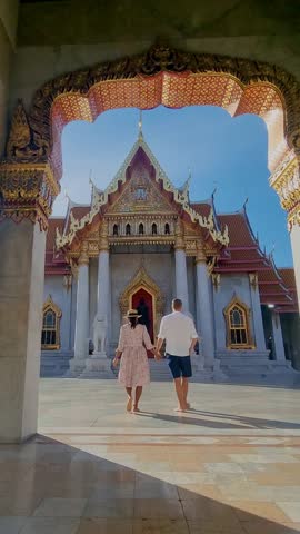 A couple strolls hand in hand through the majestic Wat Benchamabophit in Bangkok, admiring the intricate architecture and vibrant colors under the warm sun. Perfect for travelers seeking inspiration.