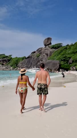 Two travelers enjoy a romantic walk along the white sandy shores of Similan Islands, surrounded by turquoise waters and lush greenery. A perfect tropical escape awaits in Thailand.