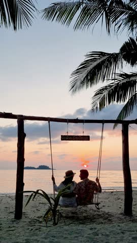 Enjoy a peaceful moment as a couple relaxes on a beach swing while the sun sets over the water on Koh Chang, Thailand. The calm atmosphere and stunning scenery create a perfect holiday escape.