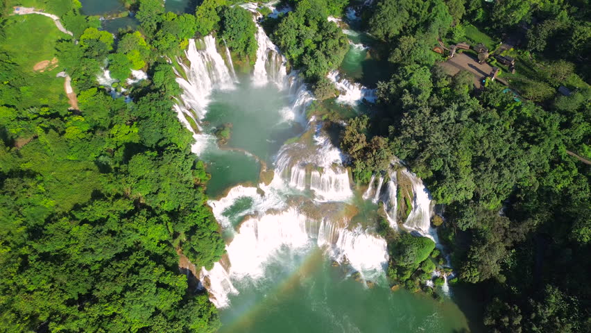 Breath-taking drone shot of the multi-level Ban Gioc Waterfall, featuring a visible rainbow over the turquoise water and small boats navigating the river.
