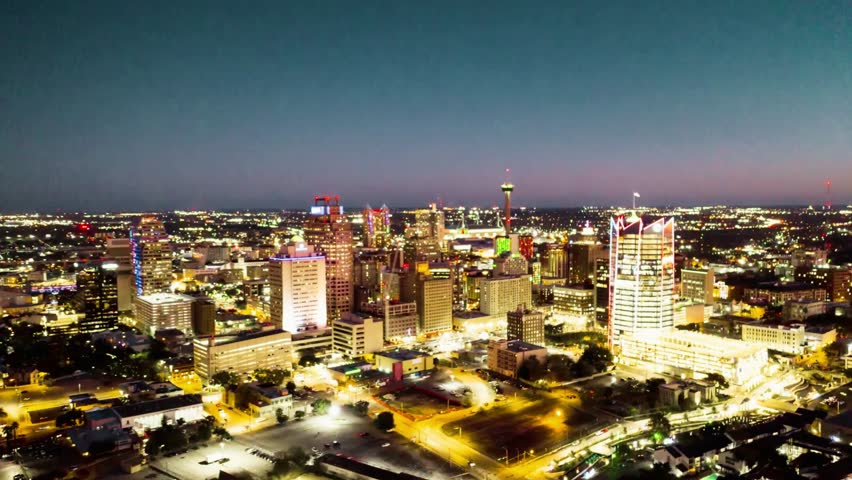 Aerial night time-lapse of San Antonio, Texas, showcasing modern office buildings and skyscrapers illuminated against the dark sky. Dynamic urban cityscape with glowing city lights, highlighting the contemporary architecture of this metropolitan area.