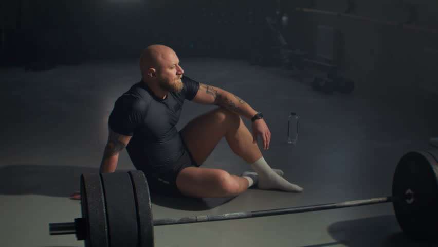 Thoughtful baldhead sportsman sits near heavy barbell on gym floor. Professional bodybuilder rests between exercises in modern sports center. Strong body