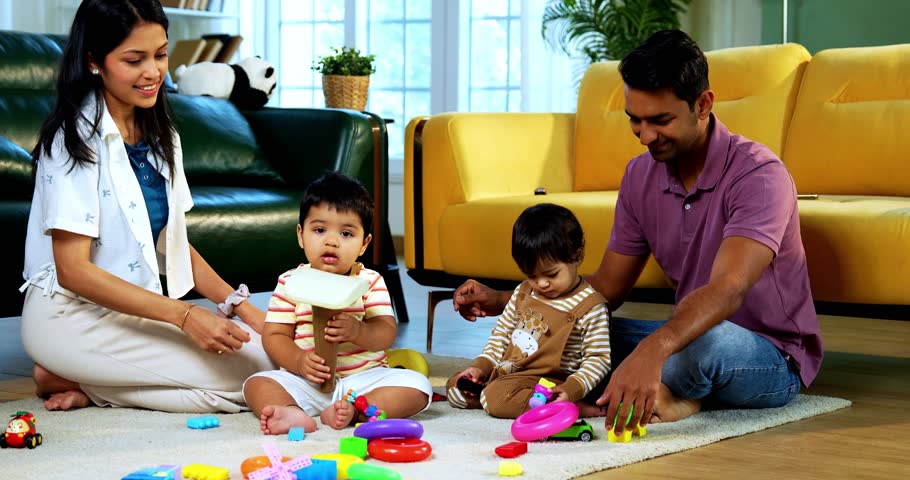 Indian Asian twin infant babies playing with colorful toys while young father and mother sit together on carpet in modern living room at home, capturing joyful family bonding and parenthood moments