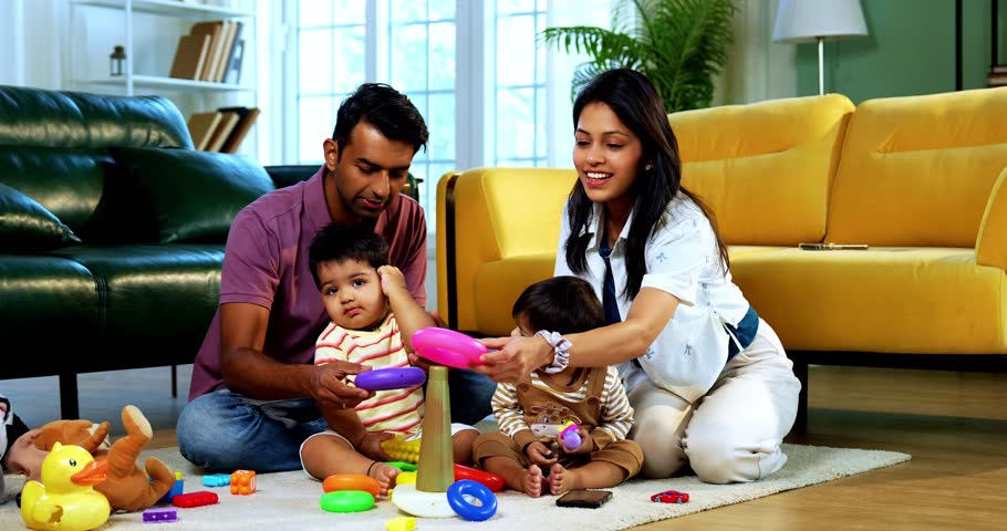 Indian Asian young parents playing with twin infant babies using colorful ring stacking toy at home, guiding fine motor learning activity and clapping with joy after completing playful task together