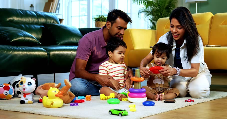 Indian Asian young parents playing with twin infant babies using colorful ring stacking toy at home, guiding fine motor learning activity and clapping with joy after completing playful task together