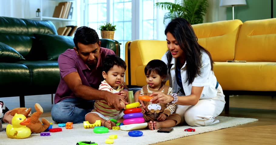 Indian Asian young parents playing with twin infant babies using colorful ring stacking toy at home, guiding fine motor learning activity and clapping with joy after completing playful task together