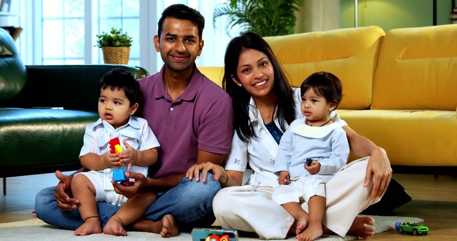 Indian Asian twin infant babies smiling and looking at camera while sitting with young parents on carpet surrounded by toys in modern home living room, capturing joyful family portrait and bonding