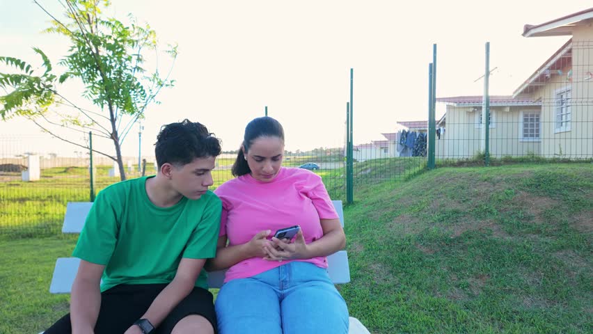 A young man and woman are sitting on a bench in a park, looking at a cell phone