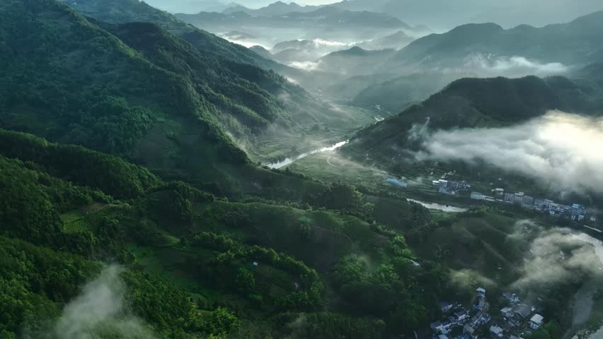 Misty mountain valley landscape with river winding through forested slopes and morning fog rolling over distant peaks and settlements.
