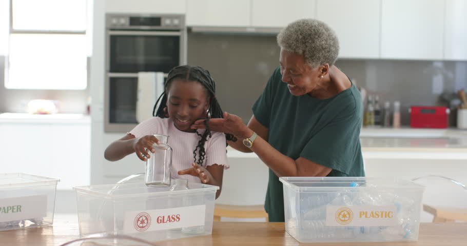 African senior woman guiding youth girl picking, placing jar into GLASS bin in kitchen, high-fiving. Recycling, grandparent, grandchild, multigenerational, mentoring, collaboration, modern