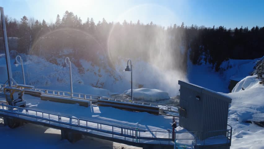 A spectacular winter landscape showing a partially frozen waterfall and river plunging into the ice, a hydroelectric dam footbridge, and mist illuminated by sunlight. Mitis-1 Power Station, Price, Quebec, Canada.