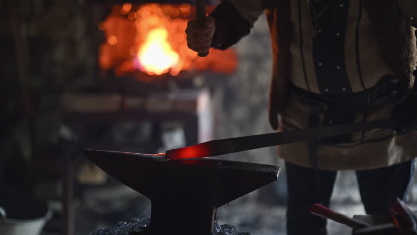 A Viking blacksmith with a white beard and braided hair works intently at a forge, hammering glowing metal on an anvil, with flames flickering in the background of a stone workshop cinematic slow
