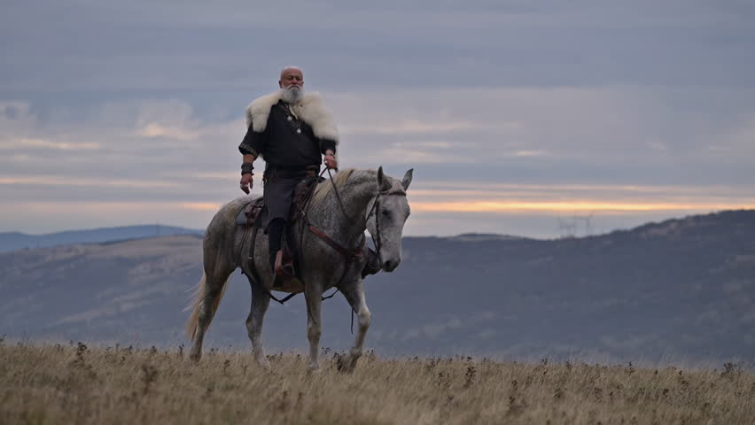 Elderly Viking warrior in fur cloak riding gray horse across open field at sunrise, mountains and distant cross behind him, moody cinematic Norse atmosphere cinematic slow motion 