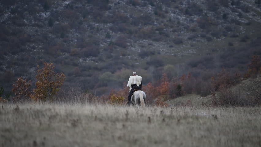 Elderly Viking warrior in fur cloak riding gray horse across open field at sunrise, mountains and distant cross behind him, moody cinematic Norse atmosphere cinematic slow motion 