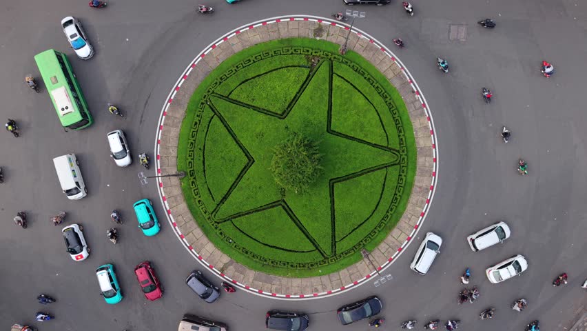 Aerial top-down view of busy roundabout in Ho Chi Minh City, Vietnam. Continuous scooter and car traffic circling green star-shaped island, dynamic urban transportation flow in Southeast Asia.