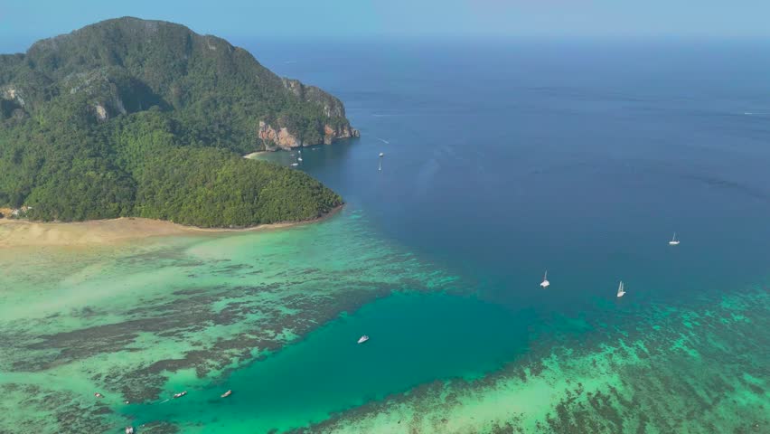 Aerial perspective reveals a stunning tropical island bay. Many colorful boats dot the clear turquoise ocean near a white sand beach.