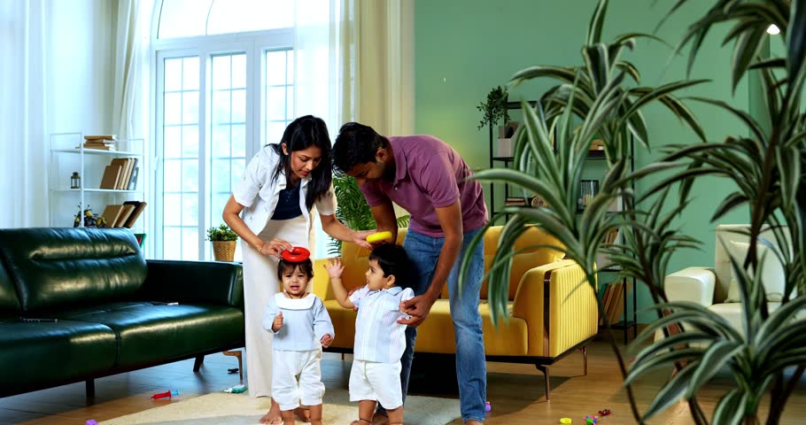 Indian Asian young parents helping twin infant babies walk by holding their hands inside modern home living room, guiding first steps together as smiling babies enjoy playful family bonding moment