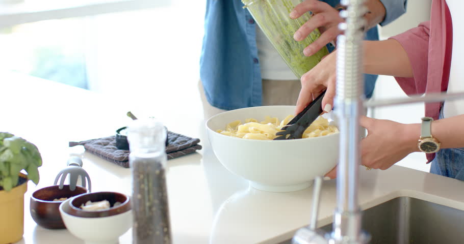 Two hands are scraping pesto from jar and are tossing pasta on white counter, coating noodles. Couple, kitchen, sauce, basil, greens, island, bright