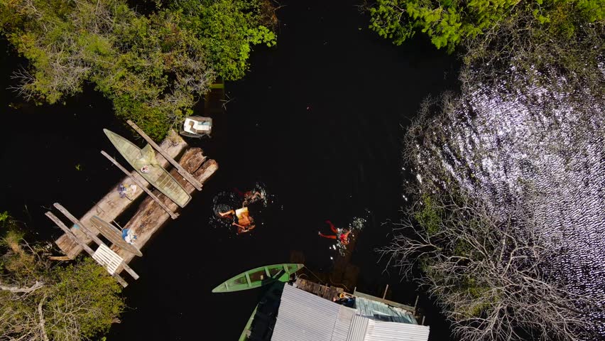 Top-down aerial of three indigenous individuals bathing in the dark Rio Negro waters. Their floating house is secluded by thick green jungle, making a high color contrast.