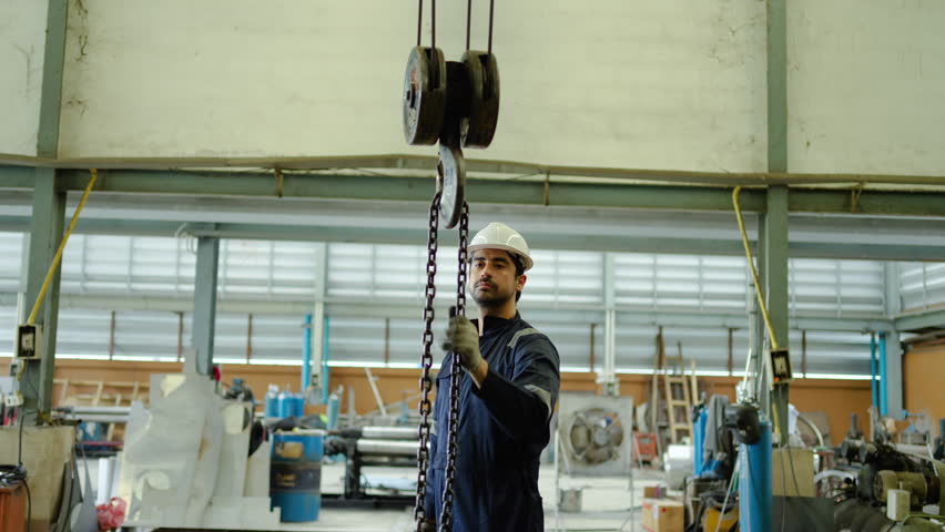 Male industrial worker wearing hard hat and safety glasses pulling heavy metal chain on overhead crane hoist in manufacturing plant. Concept of factory labor, heavy lifting, and warehouse logistics.