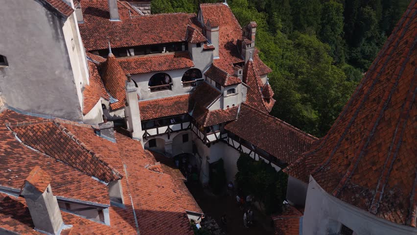 Drone shot pulling back and rising above the courtyard of Bran Castle, revealing red tiled rooftops, towers, and surrounding forest.