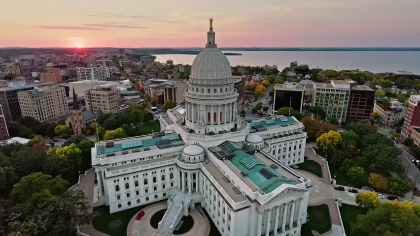 Aerial sunrise view of the Wisconsin State Capitol in Madison, WI, with the iconic dome glowing above the city skyline and surrounding streets.