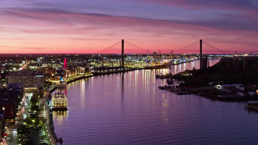 Aerial view of Savannah riverfront at dusk with glowing city lights and the iconic Talmadge Memorial Bridge spanning the water.