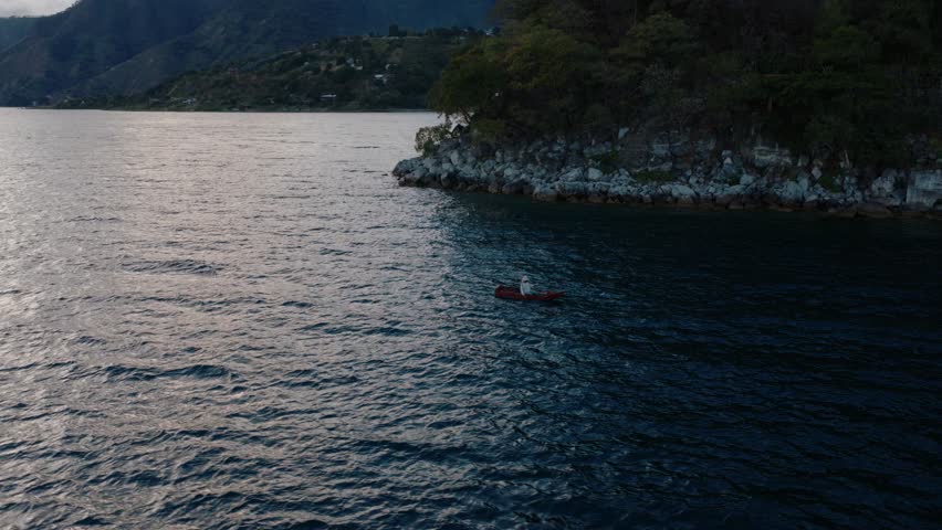 A small canoe drifts across Lake Atitlan near a rocky shoreline at dusk