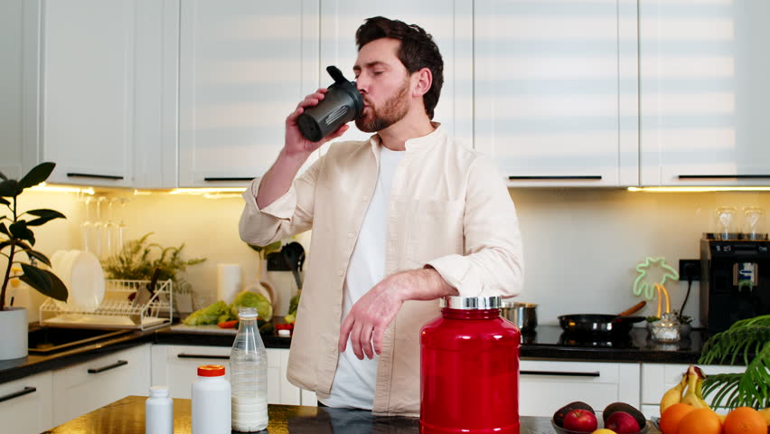 Middle-aged man smiling while drinking protein shake from shaker in home kitchen after gym workout for muscle recovery. Young guy follows diet plan for fitness training using sports nutrition at home.
