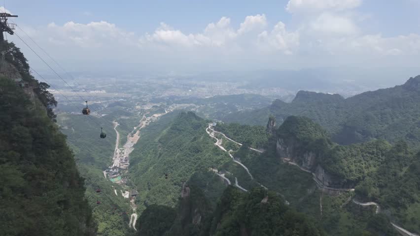 Wide angle view of cable cars gliding over the lush green peaks of Tianmen Mountain in Zhangjiajie, featuring the famous winding road and valley below. Perfect for travel and tourism content.