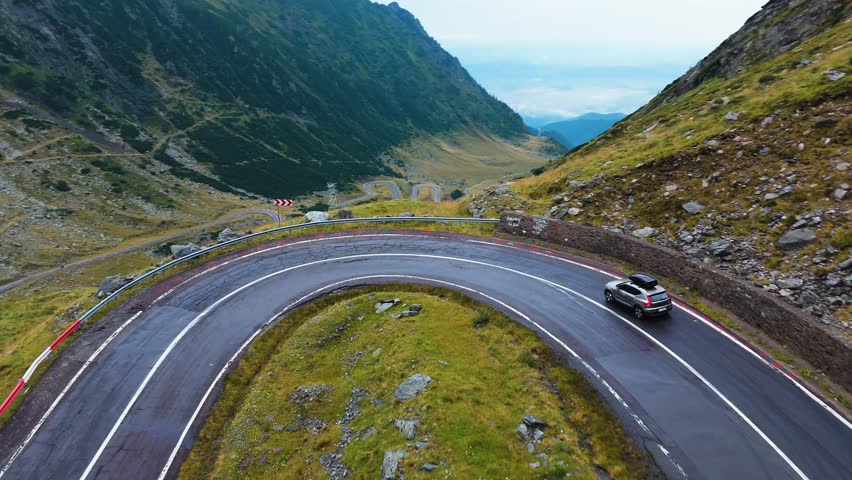 Drone shot following a car descending a tight hairpin bend, revealing the winding Transfagarasan mountain road and the deep valley below.