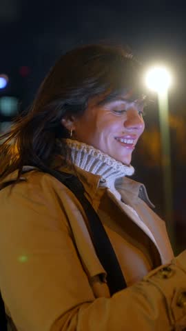 Cheerful young woman texting on her mobile phone while standing on a city street at night, with the bright screen illuminating her face and colorful bokeh lights from traffic in the background