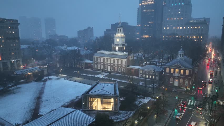 Independence Hall in Philadelphia during winter snowfall at dusk, with light traffic, glowing streetlights and historic architecture surrounded by calm blue-hour city atmosphere. Aerial wide shot.