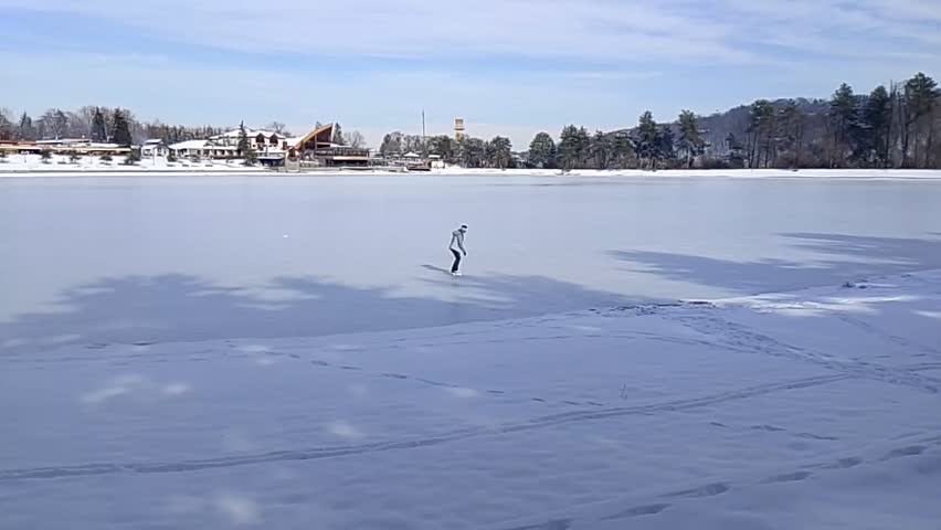A woman skates on a frozen lake in the resort area of ​​a city park, amid a picturesque winter landscape of snow-covered shores and mountainous wooded terrain.